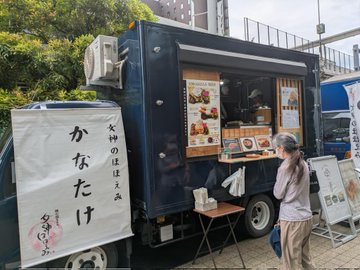 The image shows an Asian food truck parked next to a sidewalk with two people standing nearby. One person appears to be ordering from the food truck, while the other person seems to be observing or waiting for their turn. The food truck has a sign that says "Onigiri and Pork Soup" on it, indicating what is being served. There are also some Japanese characters visible in the image, suggesting that this could be an Asian restaurant or food truck serving traditional Japanese cuisine.