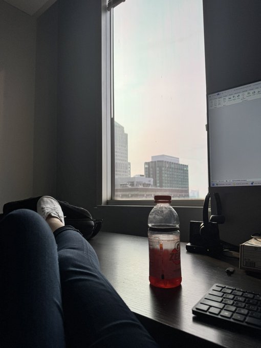 A first-person view of someone resting their legs on a desk next to a window overlooking a city, with a half-empty bottle of pink soda, a keyboard, and a monitor visible.