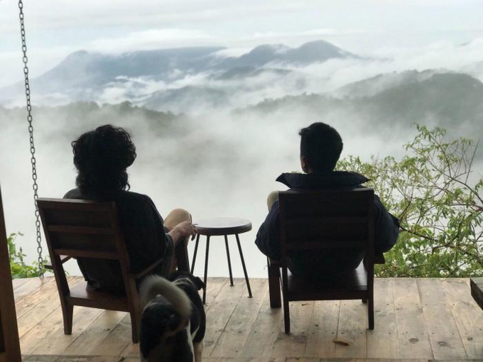 Two people sit in wooden chairs on a deck overlooking a misty mountain valley, with a black and white dog standing between them.