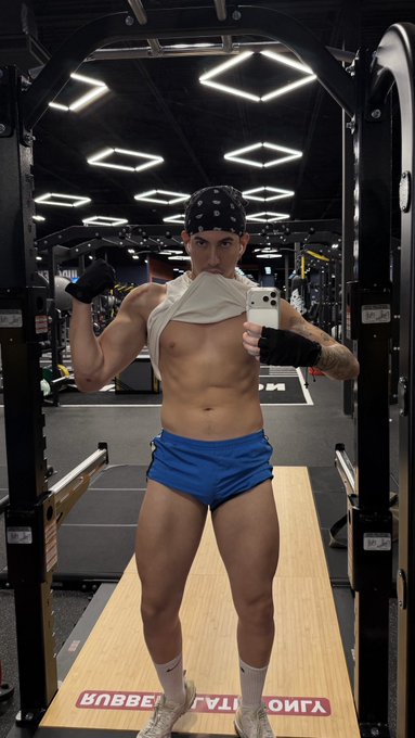 A muscular man wearing a bandana, blue boxer briefs, and fingerless gloves poses shirtless in a gym, holding a white smartphone to take a mirror selfie.