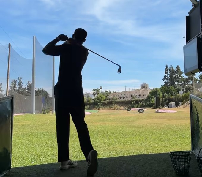 A silhouetted golfer follows through on a swing at an outdoor driving range, with distance markers "25," "50," and "70" visible on the fairway, capturing a focused practice session at Nomisma Network.