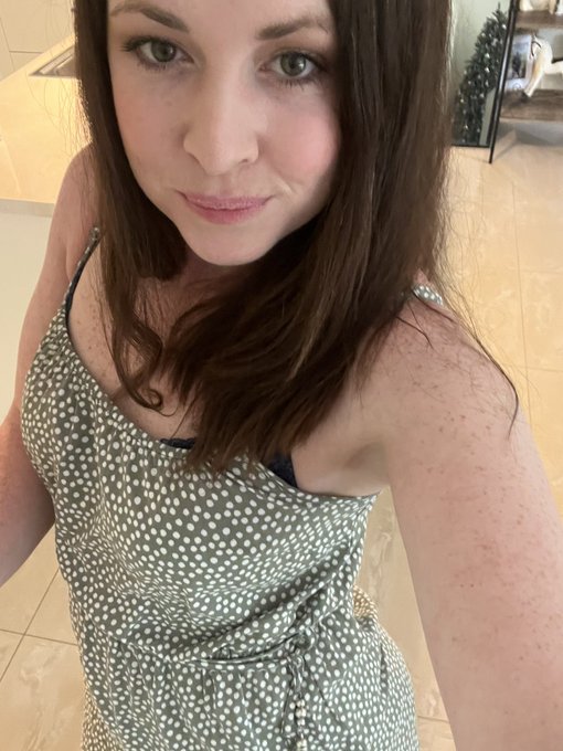 A woman with brown hair wearing a green and white polka dot dress takes a close-up selfie indoors.