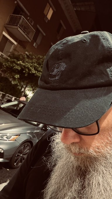 A close-up nighttime selfie of a bearded man wearing glasses and a dark baseball cap, taken on a street with a parked silver car and an apartment building visible in the background.