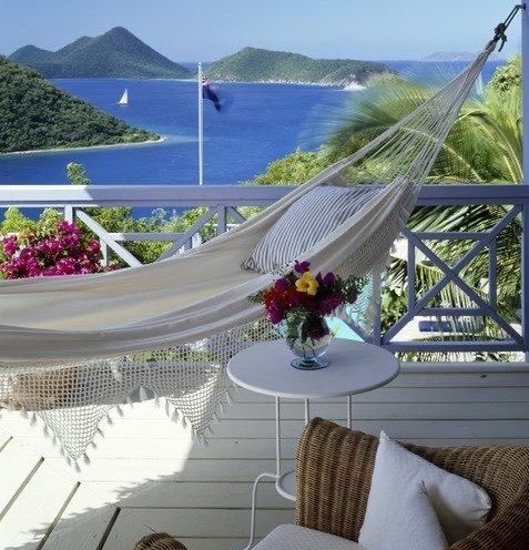 A relaxing balcony scene featuring a white hammock, a small table with a vase of colorful flowers, and a stunning ocean view with green mountains and sailboats in the distance.