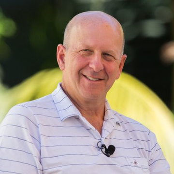 A portrait of a smiling bald man wearing a white striped polo shirt with a small lapel microphone, set against a blurred green background.