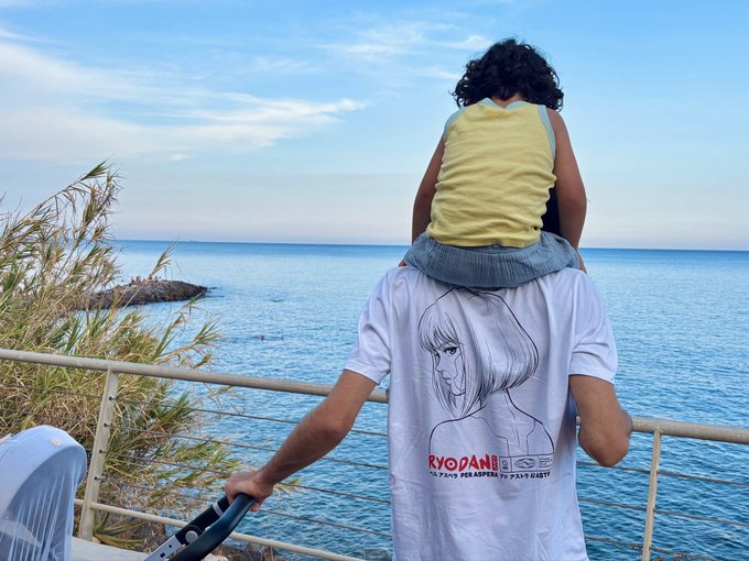 A person with dark hair wearing a yellow top sits on the shoulders of someone wearing a white 'RYO DANI' t-shirt, looking out at the sea from a seaside railing.