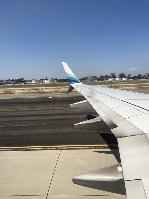 A view from an airplane window shows the wing extending over a runway with buildings in the distance under a clear blue sky.