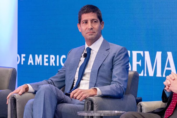 The image features a man sitting in a chair with his hands resting on his knees. He is wearing a suit and tie, giving him a professional appearance. The background of the image shows a blue wall with white text that reads "BIG: New FED Chair Kevin Warsh said Bitcoin is an important asset, and it doesn't trouble me." This suggests that the man might be participating in a panel discussion or event related to cryptocurrency, as indicated by the reference to Bitcoin.