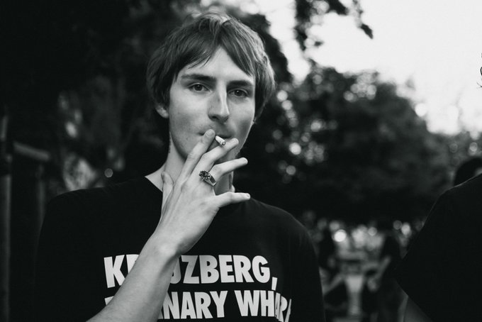 Same black and white portrait as image 2 - a young man smoking a cigarette while wearing a Kreuzberg, Germany themed t-shirt and a decorative ring.