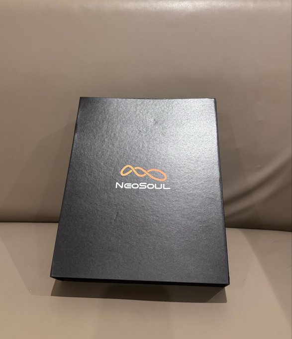 A matte black square box with the gold "NeoSoul" infinity logo printed on top, sitting on a beige surface.