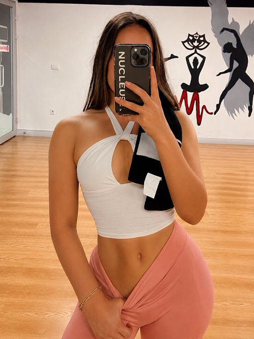 A young woman in a white halter crop top and pink leggings takes a mirror selfie in what appears to be a gym, with a 'NUCLEUS' phone case visible and a dance-themed mural in the background.