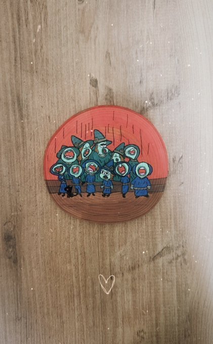 A small circular pin or sticker featuring cartoon characters with blue hoods against an orange sun background, with a small heart drawn below it on a wooden surface.