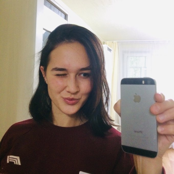 A young woman winks at the camera while holding up a silver iPhone (with the Apple logo and '16GB' visible on the back) for a mirror selfie, wearing a maroon sweatshirt in a room with bright window light.