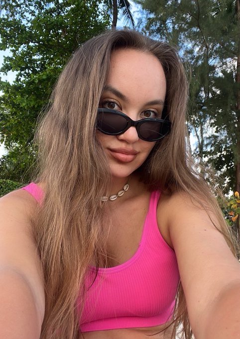 A woman with long brown hair takes a selfie outdoors wearing dark oval sunglasses, a pink sports bra, and a shell choker necklace.