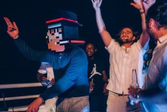 A group of people at a party, with one person wearing a pixelated Minecraft-style block head, while others raise their hands and hold drinks.