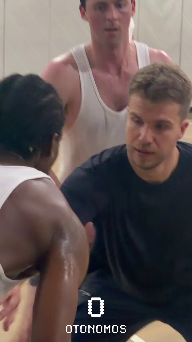 The image shows three men playing basketball together, with one man sitting down while the other two stand around him. The background of the photo features a white wall that has the word "otonomos" written on it.