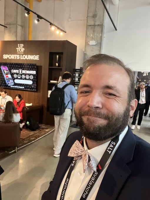 A bearded man wearing a sparkly bow tie and a lanyard with 'SALT' text takes a selfie in a convention hall, with an 'UP TOP SPORTS LOUNGE' sign and 'CAVS REWARDS' display visible behind him.
