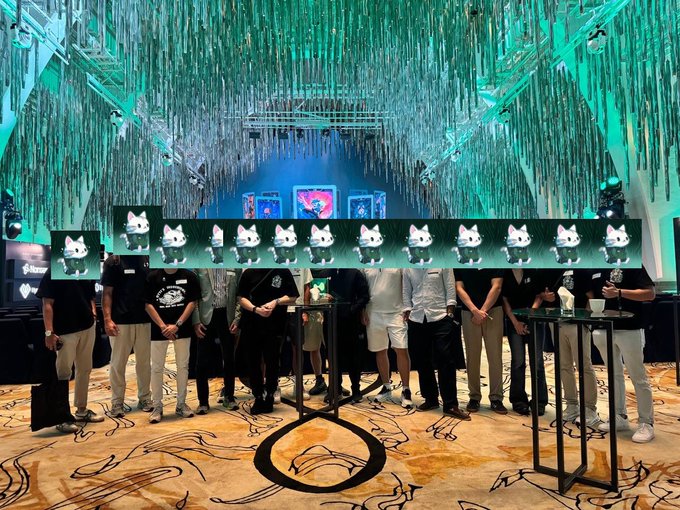 A group of people stand together at an indoor event venue decorated with green cascading streamers and screens displaying animated cat logos, with a "Hypurr" branded stand visible. The image aligns with the post celebrating HypurrCo's gathering of community leads and regional communities, showcasing the team's appreciation for the organizers behind the event.