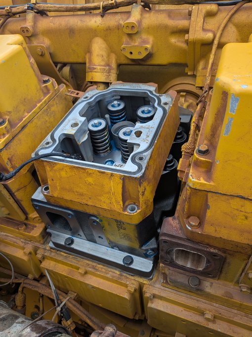 A close-up view of heavy machinery components shows a yellow-painted engine or pump housing with its cover removed, revealing internal springs and valve mechanisms.