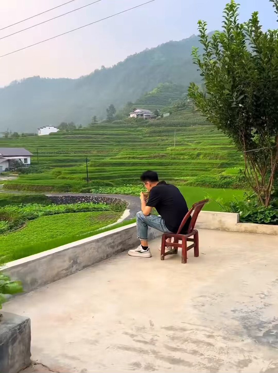 The image features a man sitting alone on a chair, looking out over a large field with mountains in the background. The scene appears to be captured from an elevated position, possibly at the top of a hill or a rooftop. The man seems to be enjoying his time and taking in the beautiful view of the landscape below him.