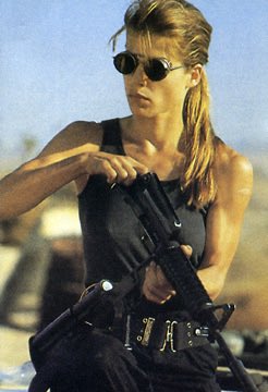 A blonde woman wearing sunglasses and a black tank top holds a tactical rifle in an outdoor desert-like setting.