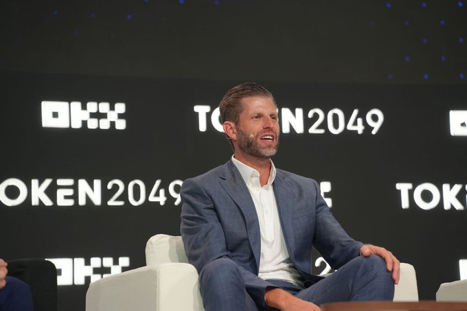 Eric Trump, seated on stage in a navy suit at TOKEN2049, smiles during the conference as the backdrop displays the event's branding and "OKX" logos. The image captures him mid-presentation, aligning with the post's quote about stablecoin adoption and credit card companies embracing the technology.