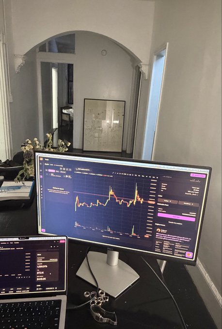 A desk setup displays a monitor showing a trading chart with colorful spikes and a laptop with financial data, with a mirror in the background reflecting a room interior.