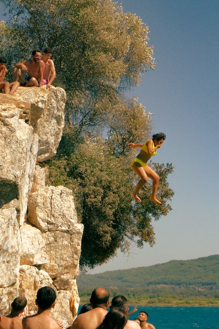 A vintage-style photograph of a woman in a yellow two-piece swimsuit mid-jump from a rocky cliff toward the water, while people watch from above and below.