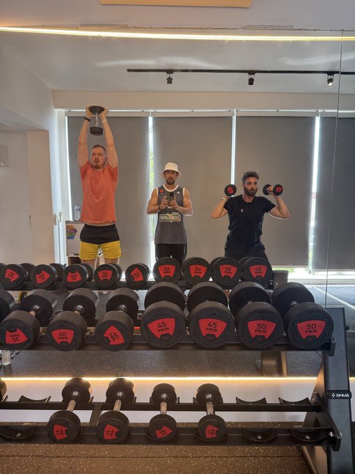 Three men lift dumbbells in a gym, reflected in a mirror above a rack of red and black dumbbells.