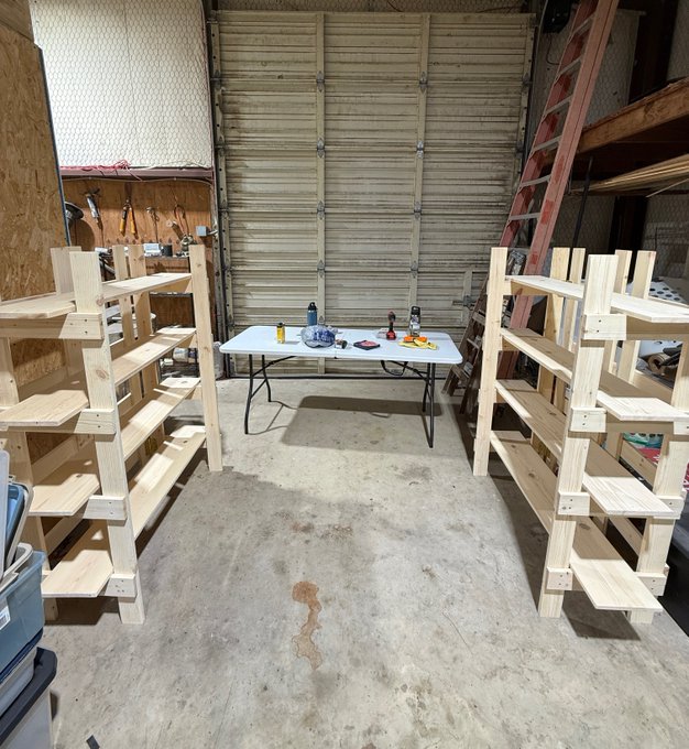 A garage interior features two tall, unfinished wooden shelving units flanking a white folding table holding tools and supplies.