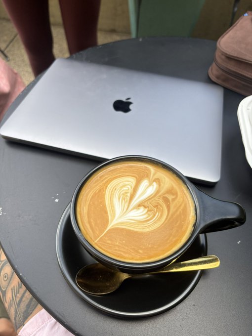 A freshly prepared latte with beautiful heart-shaped latte art sits in a black cup on a saucer with a gold spoon, positioned next to a silver MacBook laptop on a dark table at what appears to be an outdoor café.