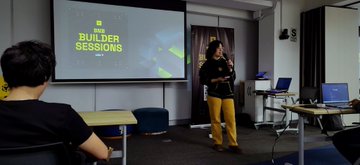 A photo from a 'Web3 Builder Sessions' event showing a woman in yellow pants presenting in front of a projection screen while an audience member watches.