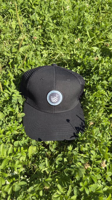 A black trucker-style cap featuring a circular white logo rests on a bed of green grass and clover.