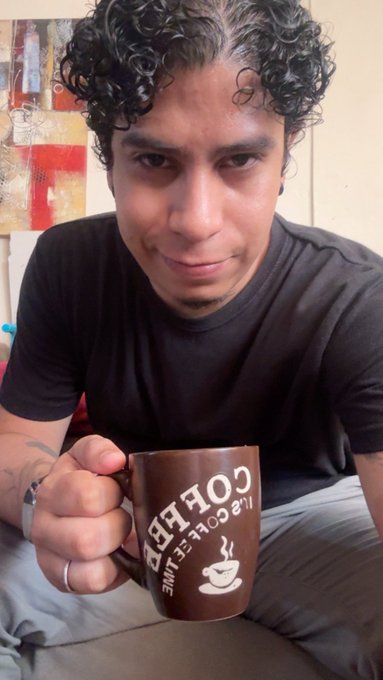 A young man with curly hair smiling while holding a brown coffee mug that reads 'COFFEE LIFE COFFEE' upside down.