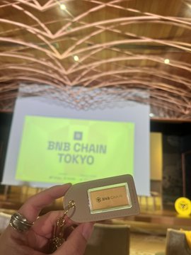 A photo of a hand holding a BNB Chain branded tag in front of a screen displaying 'BNB CHAIN TOKYO' at what appears to be a conference or event.