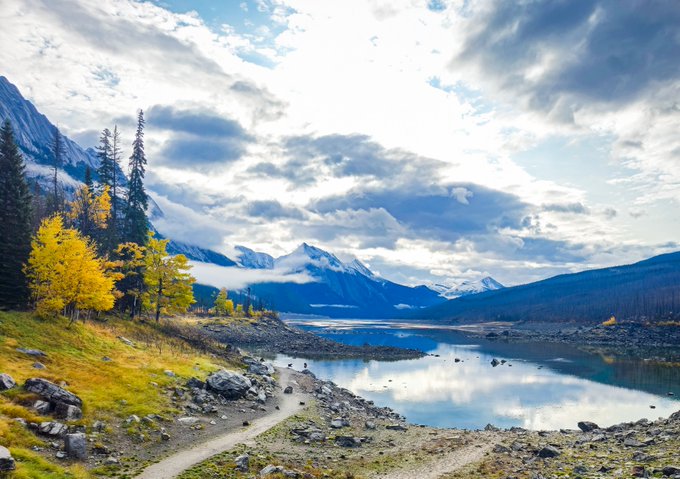 A peaceful mountain lake reflects dramatic cloudy skies as golden autumn trees frame a winding trail, capturing the serene beauty of a crisp morning greeting.