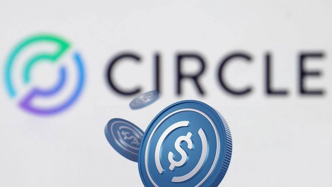 A close-up of a blue USDC stablecoin coin in front of the Circle company logo on a white background.