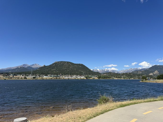 A scenic landscape photograph featuring a calm blue lake in the foreground with forested mountains and snow-capped peaks under a clear blue sky, with a paved path visible on the right.