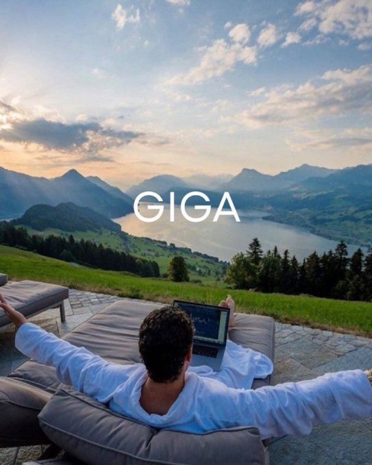 A man in a white shirt relaxing on lounge cushions with a laptop, overlooking a scenic mountain lake landscape, with the text 'GIGA' overlaid in the center.