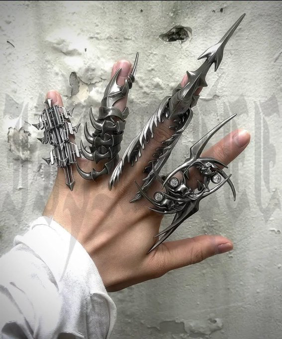 A hand displaying an elaborate set of dark metallic gothic-style rings and claws with intricate pointed, skeletal, and demonic designs.