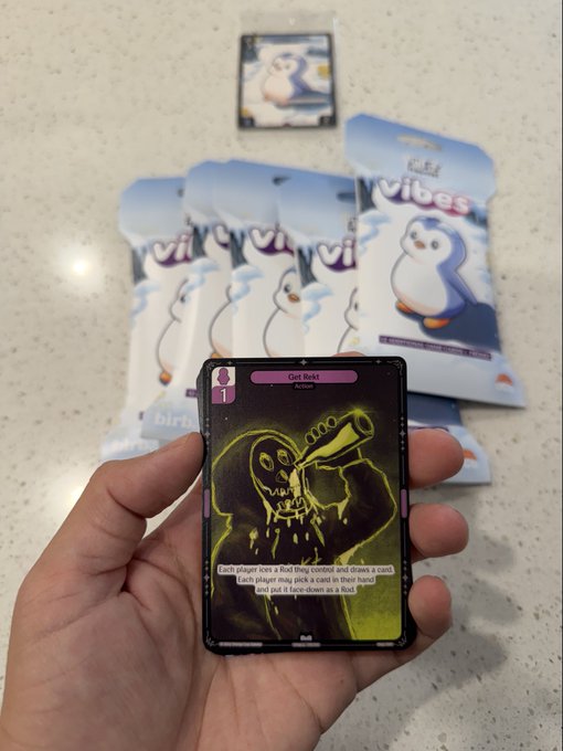 The image features a hand holding up a small card with a cartoon penguin on it, along with some text that reads "Pulled a @rektguy @vibes_tcg!". The person's fingers are visible as they hold the card in their hand. In the background, there is a collection of Vibes cards and a penguin-themed game box, suggesting that this image might be related to a gaming event or convention where players can try out different games and collect cards with various characters from the games.