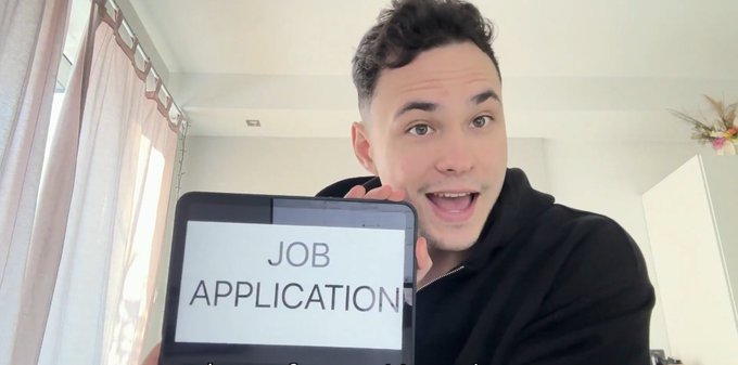 The image features a man holding up a sign that reads "Job Application", possibly for an online job posting or a real-life event. The background of the image shows a window, suggesting that they are indoors. The text on the sign is in black, making it stand out against the white background. The man's facial expression and body language convey excitement about the opportunity to apply for the job.