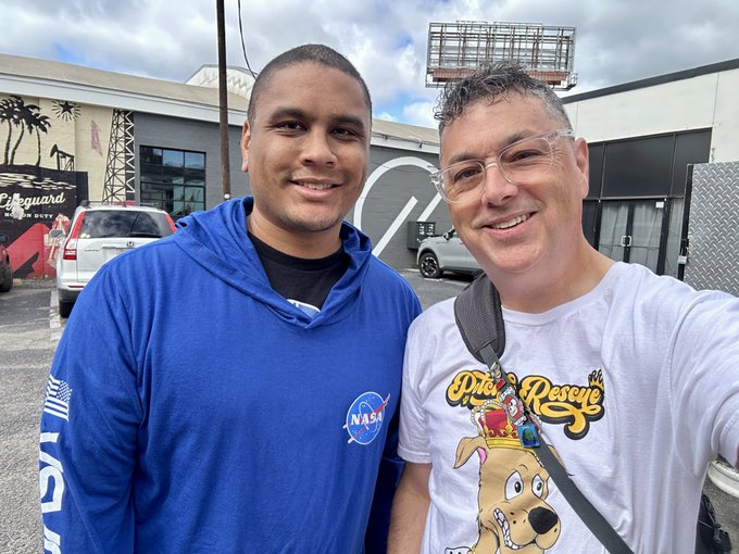 A selfie of two men smiling outdoors; the younger man wears a blue NASA hoodie, while the older man wears a white graphic t-shirt, with buildings and vehicles in the background.