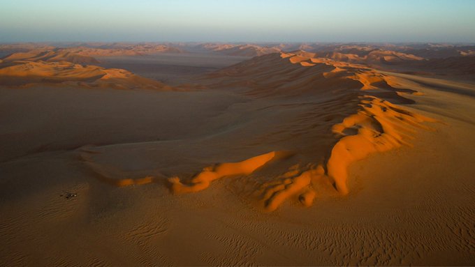 An aerial view shows vast sand dunes stretching across a desert landscape under a clear sky, likely at sunset or sunrise.