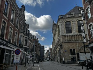 A European city street scene featuring traditional architecture with a 'HOTEI DEBROUWER' hotel building on the right under a partly cloudy blue sky.