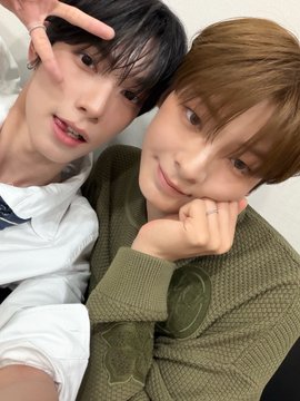 The image features two young men posing for a picture, both wearing green sweaters. They are sitting next to each other with their hands resting on their laps or behind their backs. The background of the photo appears to be a wall, providing a simple backdrop that allows the focus to remain on the two boys and their interaction.