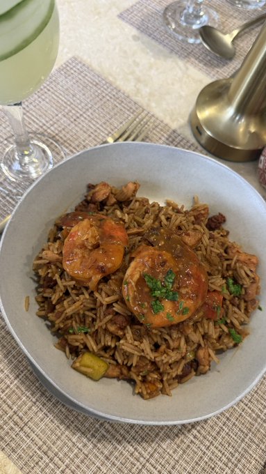 A plate of seasoned rice with two pieces of shrimp in red sauce garnished with parsley, served on a placemat alongside a wine glass and lamp.