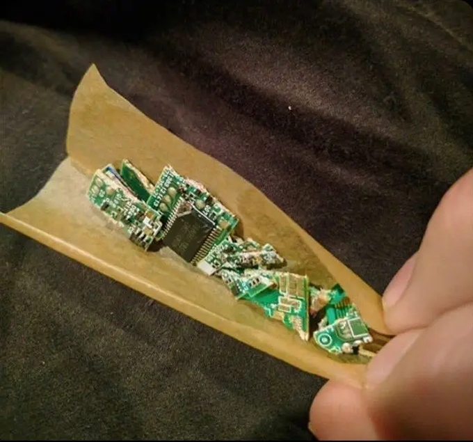 A person's hand holding a small piece of paper or envelope containing several small pieces of broken green circuit boards with electronic components and a microchip.