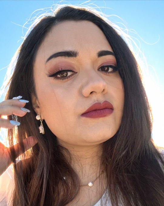 A close-up portrait of a woman with long dark hair, dramatic eye makeup, mauve lipstick, and a daisy drop earring, taken outdoors against a blue sky.