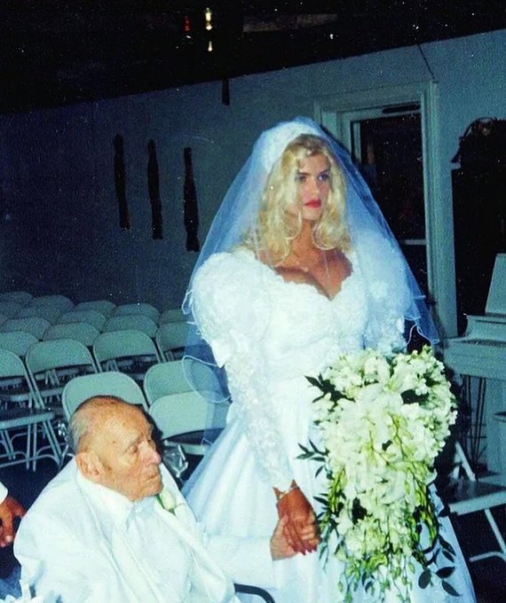 A blonde bride in a white gown and veil holds a large bouquet of white flowers while standing beside a seated elderly man in a white shirt, in what appears to be a wedding ceremony venue with empty chairs in the background.
